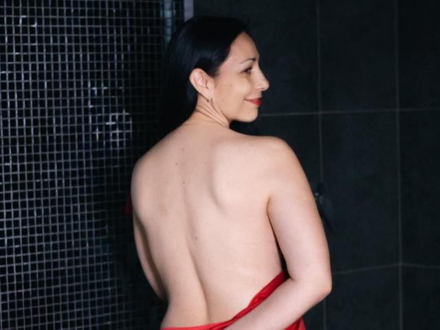 LiluGrants with black hair, bare back, and red cloth, showing off her ass, smiling, in a dark-tiled bathroom, looking over her shoulder.