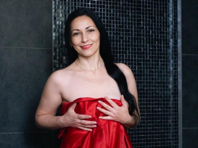 Lilu, com longos cabelos pretos, vestindo um vestido de cetim vermelho, cobrindo os seios com as mãos, sorrindo, em pé em um banheiro escuro com azulejos, usando um colar fino..