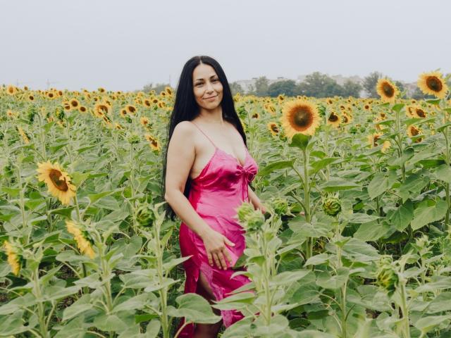 LiluGrants with long black hair, pink satin dress, small tits, standing in sunflower field, smiling, hands on hips, sunny day, green leaves, yellow sunflowers, clear sky.