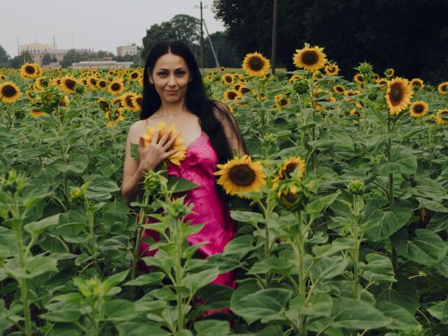 LiluGrants with long dark hair, pink dress, standing in sunflower field, holding sunflower, green leaves, yellow petals, sunny day, smiling, cute, attractive, natural light.
