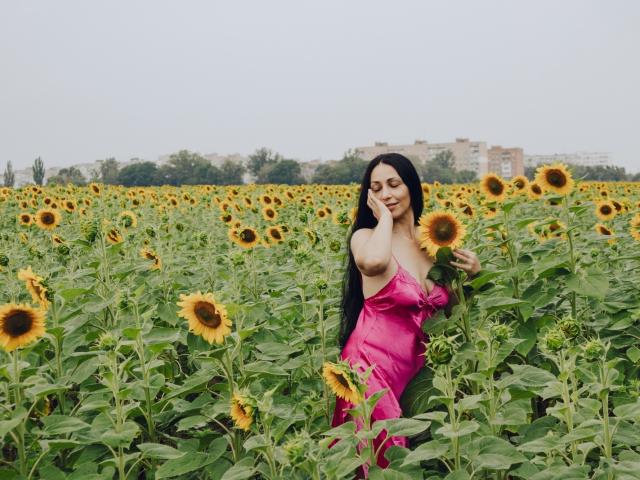 LiluGrants with long black hair, wearing a pink dress, touching her lips, standing in a sunflower field, holding a sunflower, looking cute, tits visible, sexy.