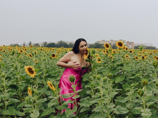 LiluGrants stands in a sunflower field, wearing a pink dress, covering her tits with one hand, surrounded by tall green plants and bright yellow sunflowers.