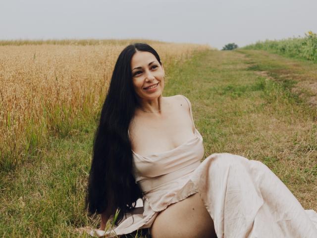LiluGrants, with long black hair, sits in a grassy field, wearing a pale pink off-shoulder dress, smiling, showing her tits and ass, in a wheat field.