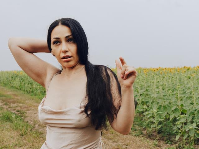 LiluGrants with long black hair, tan skin, and large tits in a beige dress, pointing with her right hand, standing in a sunflower field, looking at the camera.