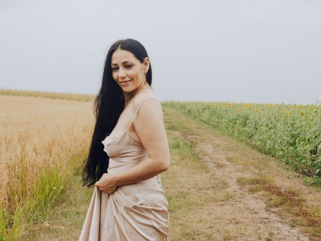 LiluGrants, met lang zwart haar, lachend, gekleed in een beige jurk, houdt iets vast, staand op een zandpad, tussen een gouden veld en een groen gewas..