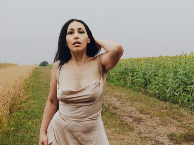 LiluGrants has big tits, dark hair, and a hand in her hair, wearing a beige dress, standing on a dirt path with sunflower fields on the right.