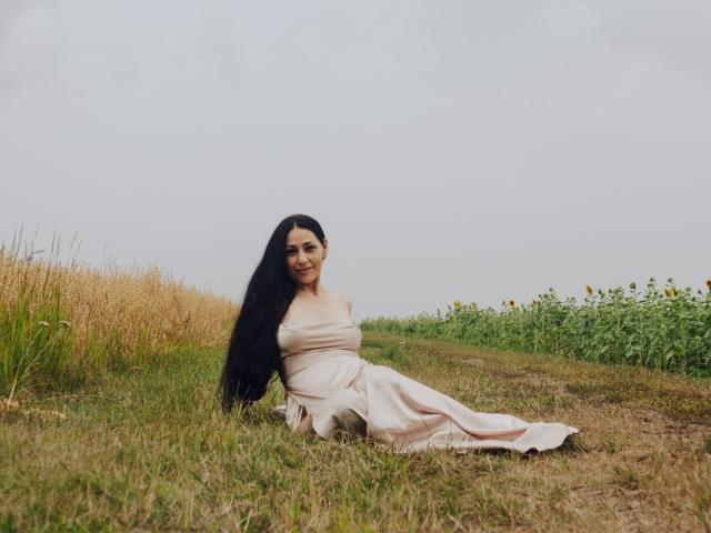 LiluGrants with long black hair, sitting on grass in a pale pink dress, smiling at the camera, showing her tits and ass, in a field with green and yellow plants.