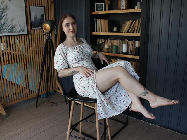 LauraHlot, pale-skinned with tattoos, sits on a black chair, legs crossed, wearing a floral dress, exposing her thighs and tattooed ankle, smiling, in a cozy room.