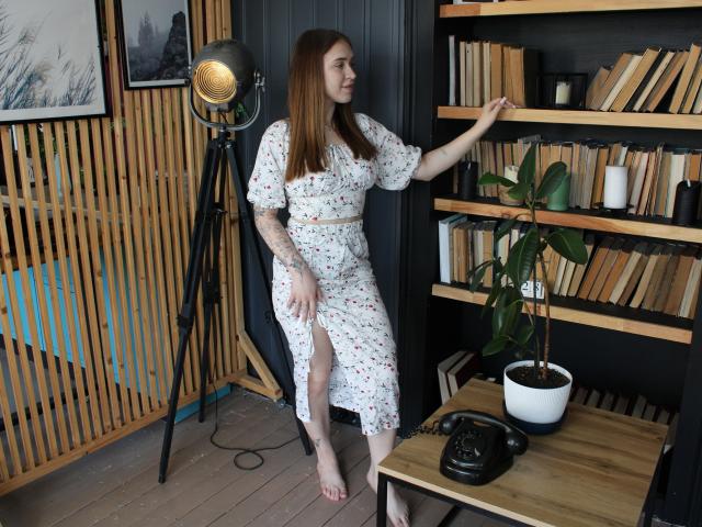 LauraHlot with long brown hair, white floral dress, and a belt, standing by a wooden bookshelf, touching the top shelf, barefoot, black vintage phone on a wooden table.