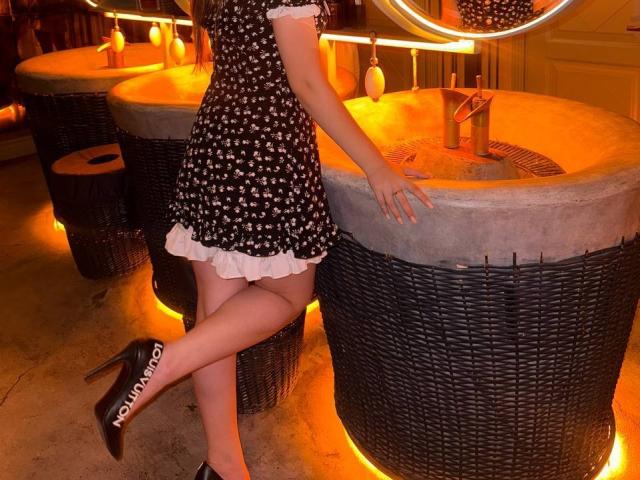 Bubochka in a black dress with white polka dots, standing with one leg crossed, wearing black high heels with "PUMPUP" on the heel, leaning on a wicker bar.