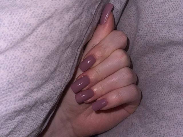 Barberry's hand with pinkish-purple glossy nails, fingers slightly curled, resting on a gray textured fabric. Skin is pale with visible knuckles. Image focuses on her hand and nails.