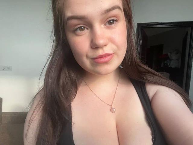 SweetVibe-hot with long brown hair, brown eyes, and pink lips, wearing a black tank top showing her big tits, and a small pink necklace, smiling at the camera.