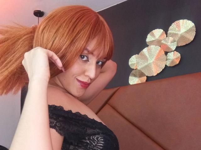KaylinKrof with red hair in a ponytail, wearing black lace, exposing her bare shoulders and smiling at the camera. Her tits are partially visible. Her hand is in her hair.