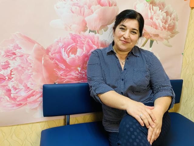 BridjetTur with medium tits, black hair, blue patterned shirt, dark polka-dot pants, sitting on blue chair, smiling, hands on knee, ring on left hand, floral wall art.