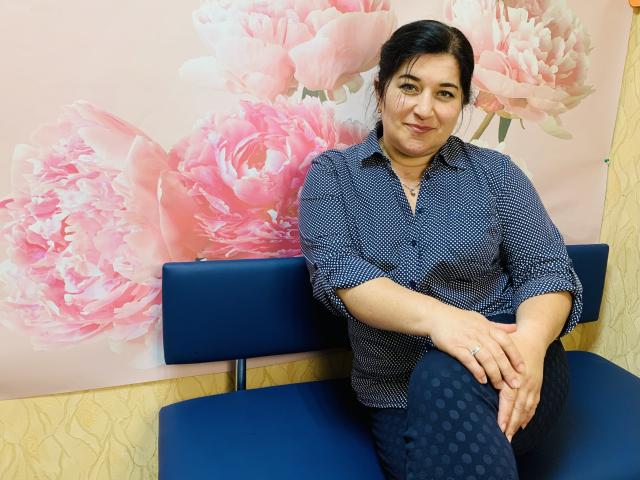 BridjetTur, chubby woman with dark hair in a blue patterned shirt and navy polka-dot pants, sitting on blue chairs, smiling slightly, hands on knees.