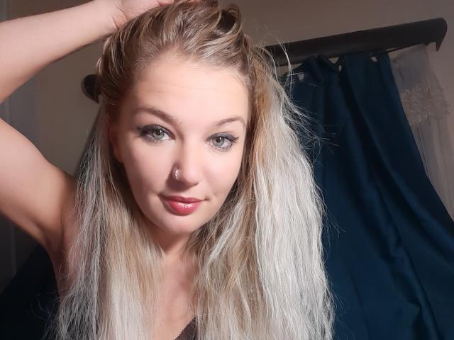 AlmaDeAngel69 has blonde hair, blue eyes, and a nose ring, with a slight smile, raising one arm, showing her tits, and a sexy look. Her pink lips are glossy.