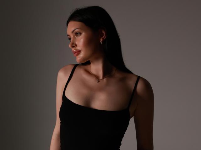 MilanaMusee with dark black hair, wearing a black tank top, showing her small boobs and slim, fair-skinned body, gazing to the side with a subtle smile.