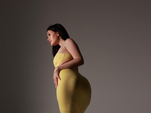 MilanaMusee with big ass, wearing a tight yellow dress, side profile, long dark hair, light skin, hands on belly, looking down, gray background.