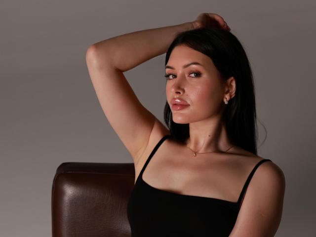 MilanaMusee with dark hair, wearing a black tank top, arms raised, showing her tits, small gold necklace, brown leather chair, subtle makeup, confident smile, soft light.