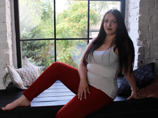 IsabelBlackberry with long black hair, white tank top, and red pants, sitting on black window ledge, legs apart, showing her ass, white brick wall, and black window frame.
