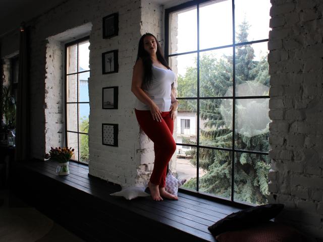 IsabelBlackberry stands barefoot on a black wooden windowsill, wearing a white tank top and red pants, with long dark hair, looking outside, her tits slightly visible.