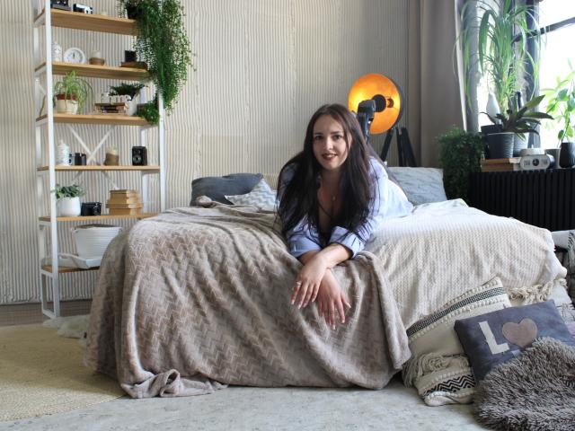 IsabelBlackberry with long brown hair, sitting on a bed with a beige blanket, wearing a blue shirt, smiling, hands on blanket, bedroom with plants and a camera.