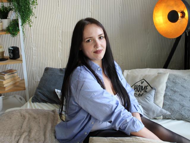 IsabelBlackberry with long black hair, wearing a blue shirt and black tights, sitting on a bed, smiling, showing small tits, with a warm light in the background.