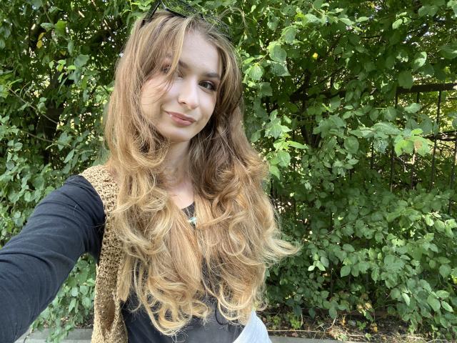 NinaNa with long, wavy blonde hair, black long-sleeve shirt, and tan crochet vest, smiling softly. Her tits are small, and she's standing in front of green leafy bushes.