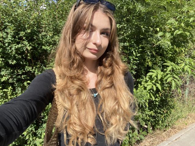 NinaNa with long, wavy, blonde-to-brown ombre hair, wearing a black long-sleeve shirt, and sunglasses on her head, standing outdoors with green foliage in the background.