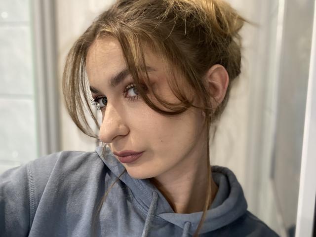 NinaNa with messy brown hair in a bun, wearing a blue hoodie, looking to the side with a slight smile, nose ring, and subtle makeup. Soft lighting.
