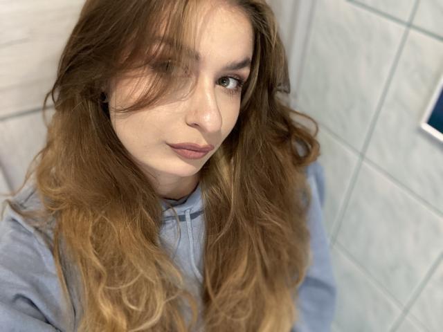 NinaNa with long brown hair, green eyes, and light makeup, wearing a gray hoodie, looking up with a slight smile, her hair partially covering her right eye.