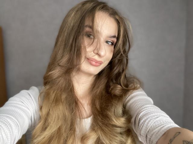 NinaNa has long, wavy brown hair with blonde highlights, wearing a white ribbed shirt, smiling with pink lips, and has a tattoo on her right arm.