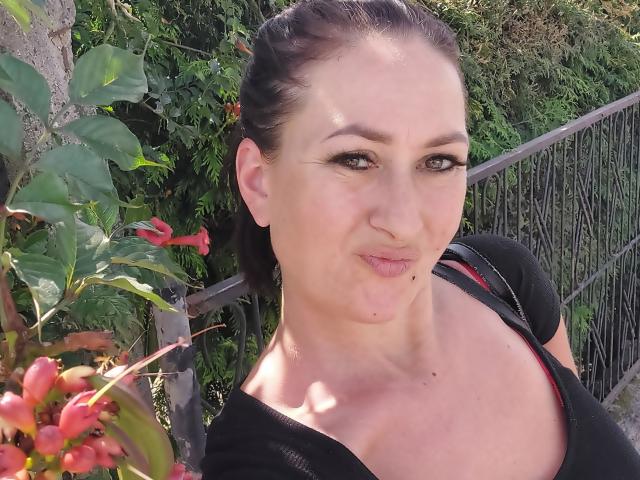 SensualNathalie with puffy lips, black hair in a ponytail, wearing a black top, showing her boobs, standing next to red flowers and green leaves, with a metal fence.