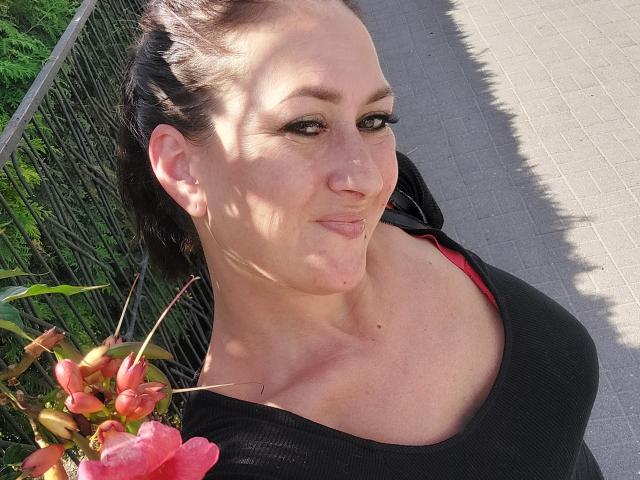 SensualNathalie has large tits, pale skin, and dark hair in a ponytail. She's wearing a black top with a red bra peeking out. Her face is slightly shadowed.