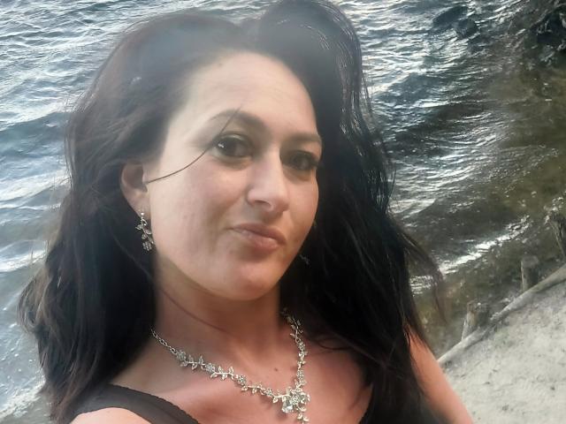 SensualNathalie with dark hair, brown eyes, and a subtle smile, wearing a silver necklace and earrings, by the water, showing a slight pout and some tan lines on her chest.