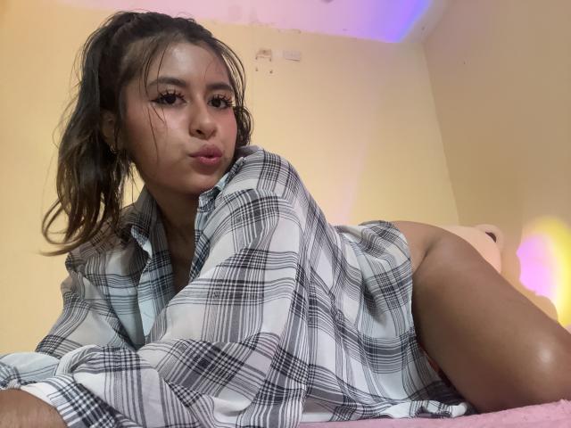 MaiaHairy with pouty lips, wearing an oversized plaid shirt, lying on bed, showing her bare ass, long eyelashes, and brown ponytail. She's cute and sexy.