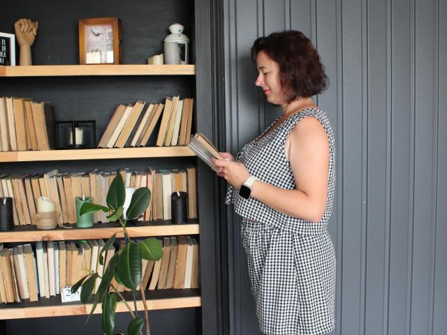MarieSho met krullend bruin haar, gekleed in een zwart-wit geruite jurk, die een boek vasthoudt, staat naast een grijze muur en houten boekenkasten met een groene plant..