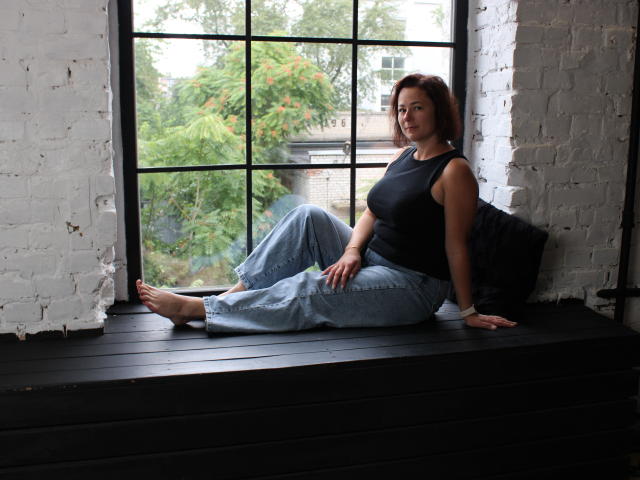 MarieSho sits on a black window ledge, barefoot, wearing a black tank top and loose blue jeans, with white brick walls and a large window behind her.