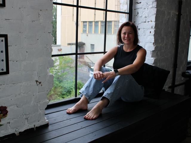 MarieSho, barefoot, sitting on black window ledge, wearing sleeveless black top, blue jeans, smiling, white brick wall, black metal window frame, wristwatch, bare toes, relaxed.