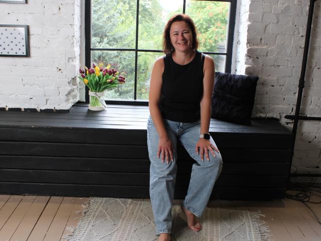 MarieSho, medium tits, black sleeveless top, blue jeans, brown hair, smiling, sitting on black wooden bench, vase of tulips, white brick wall, window, wooden floor.
