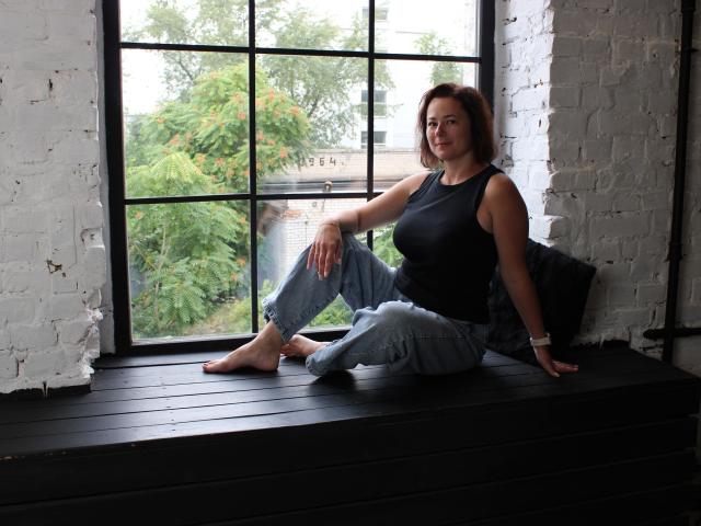 MarieSho with medium tits, wearing a black tank top and blue jeans, sitting barefoot on a black window ledge, white brick walls, and greenery outside the window.