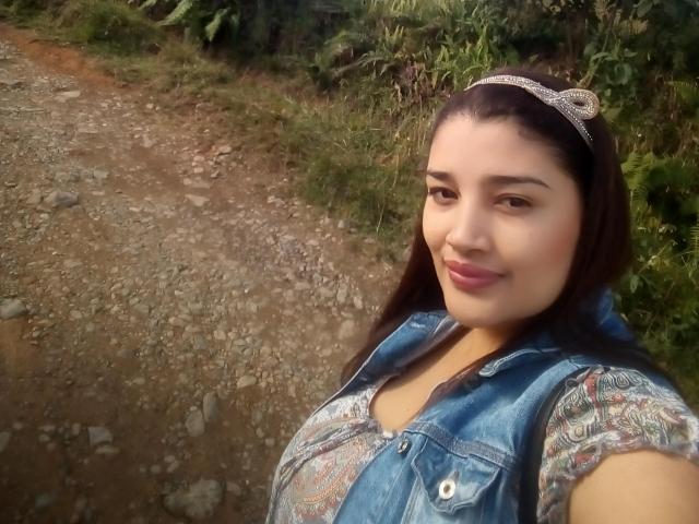 ZafiRouge with dark hair, wearing a jeweled headband, denim vest, and patterned shirt, smiling at the camera, showing her boobs slightly, standing on a rocky dirt path.