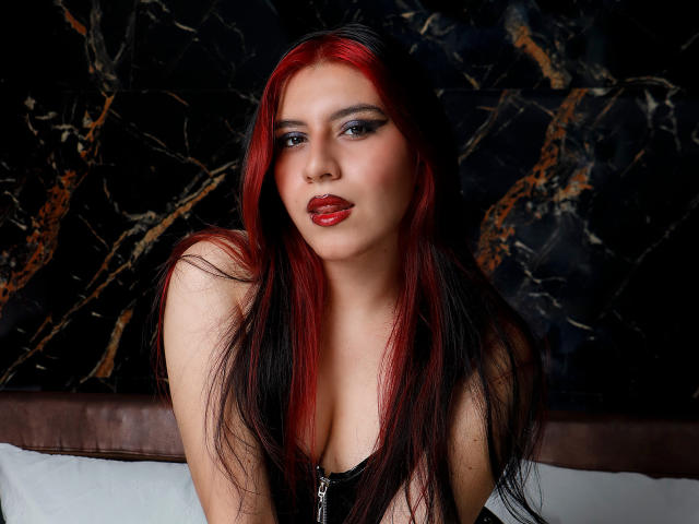 DannaOlivieri with red and black hair, wearing red lipstick and dark makeup, looking at the camera, showing her cleavage, sitting in front of a dark marble wall.