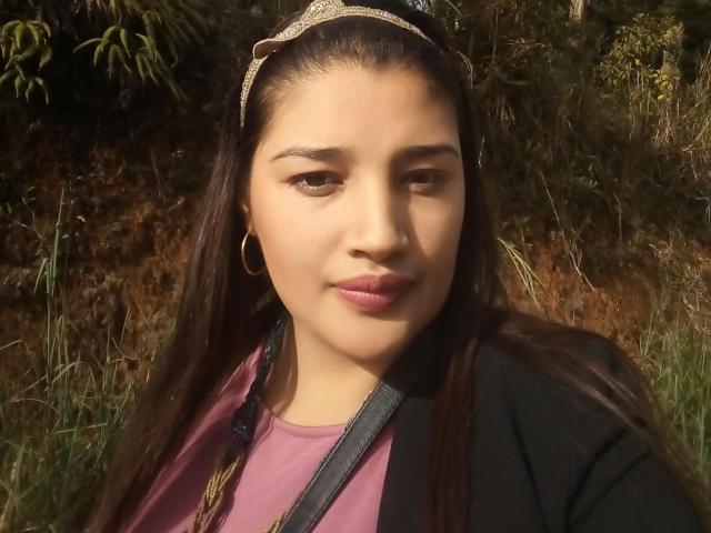 ZafiRouge with long brown hair, wearing a glittery headband, pink shirt, black jacket, and gold hoop earrings, has a subtle smile and small brown eyes. Her pussy is not visible.