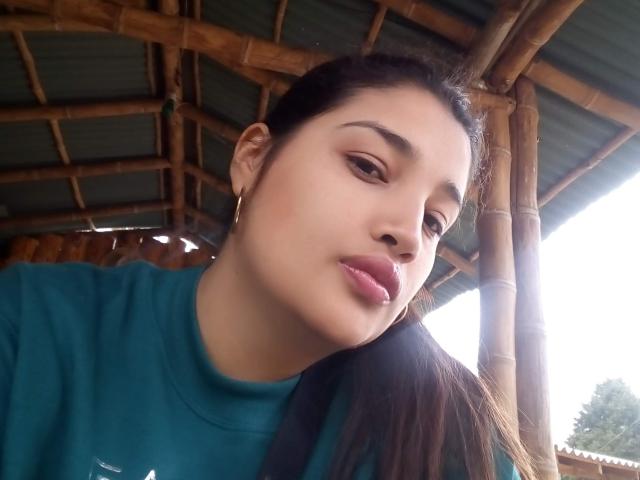 ZafiRouge with pouty lips, wearing a teal shirt, brown hair in a ponytail, gold hoop earrings, under a bamboo roof. Soft light on her face, slightly flushed cheeks.