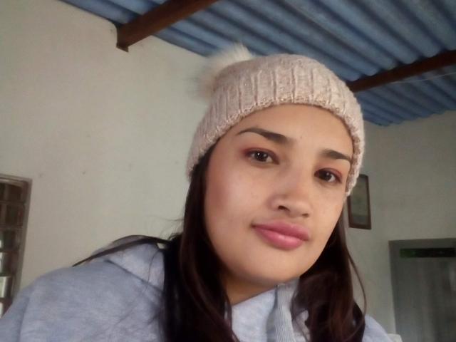 ZafiRouge, a Latina chick with dark brown hair, wearing a pink knitted beanie with a white pom-pom, gray hoodie, pink lipstick, and red eyeshadow, smiling at the camera.
