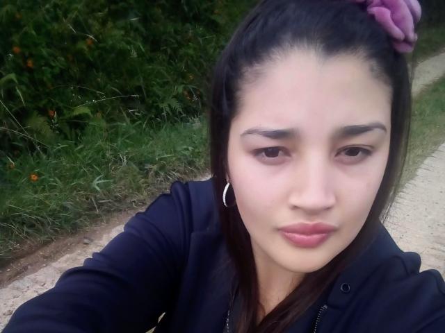 ZafiRouge has dark hair in a pink scrunchie, wearing a black hoodie, with slightly puffy lips and a neutral expression, standing on a gravel path.