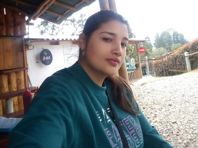 ZafiRouge has medium brown skin, dark brown eyes, and long brown hair in a ponytail, wearing a green sweatshirt, with pink lipstick, holding a camera for a selfie.
