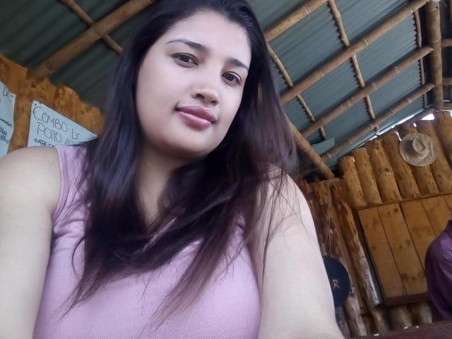 ZafiRouge with long dark brown hair, pink sleeveless top, light brown skin, subtle makeup, and a slight smile, standing in a rustic wooden building with a green roof.