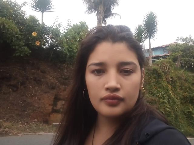 ZafiRouge has medium brown skin, long dark brown hair, and wears gold hoop earrings. She's looking straight at the camera with a neutral expression. Her small, dark pink lips are slightly pouted.
